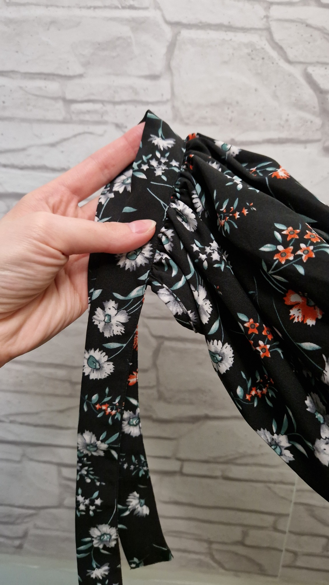 Dłoń przytrzymuje marszczony rękaw czarnej sukienki w białe i pomarańczowe kwiaty, z wiązaniem z tej samej tkaniny, na tle ściany z białej cegły.