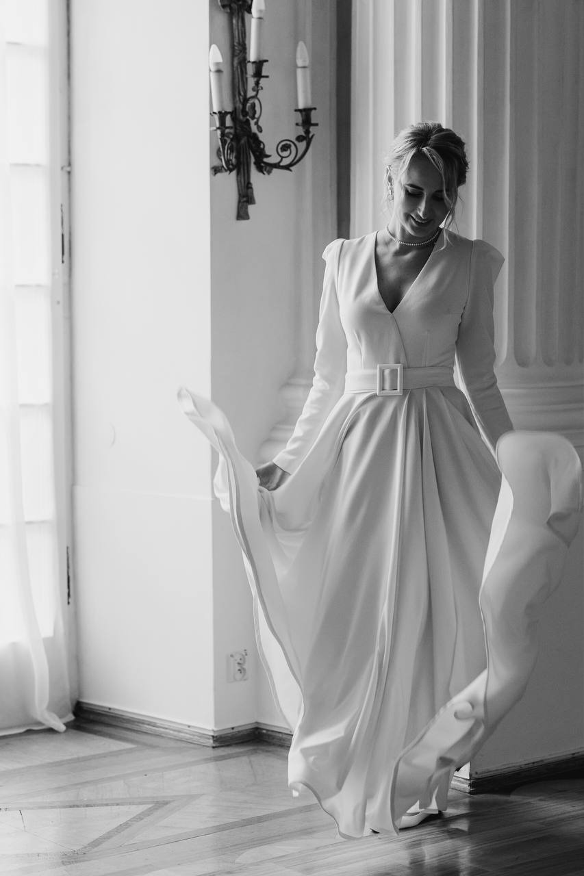 Kobieta w białej, eleganckiej sukni z paskiem, stojąca w jasnym wnętrzu z kolumnami i żyrandolem, spódnica sukni w ruchu.