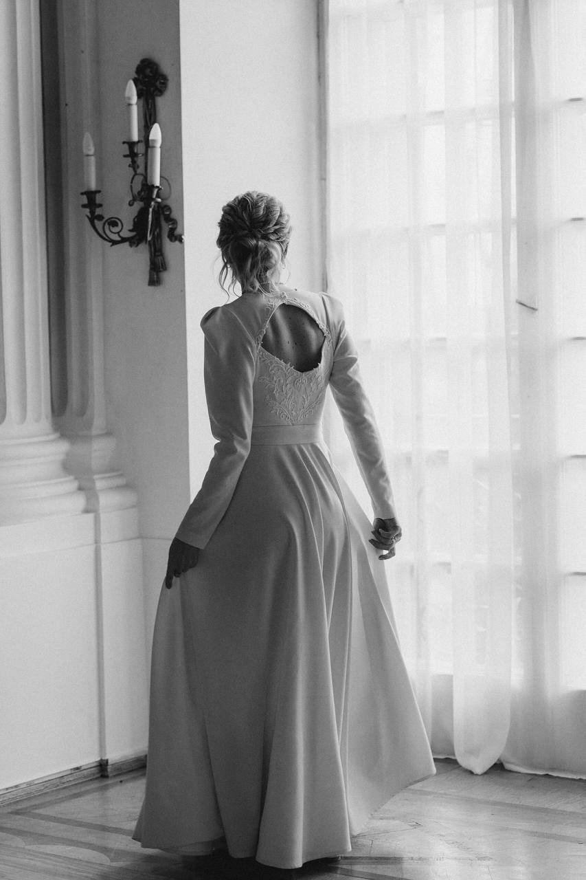 Kobieta w długiej, białej sukni z wycięciem na plecach ozdobionym koronką, stojąca tyłem w eleganckim wnętrzu z kolumnami i świecznikiem.
