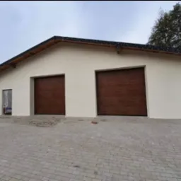 Nowo wybudowany garaż z dwiema brązowymi bramami segmentowymi i ocieploną elewacją w kolorze kremowym, otoczony kostką brukową.