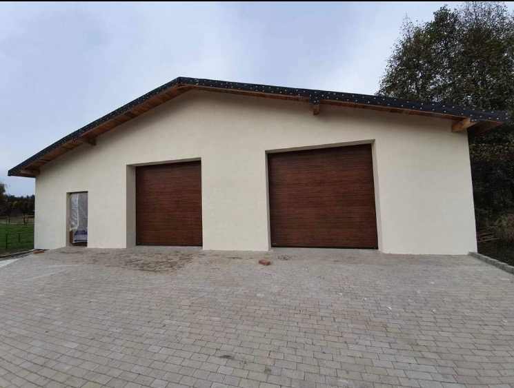 Nowo wybudowany garaż z dwiema brązowymi bramami segmentowymi i ocieploną elewacją w kolorze kremowym, otoczony kostką brukową.