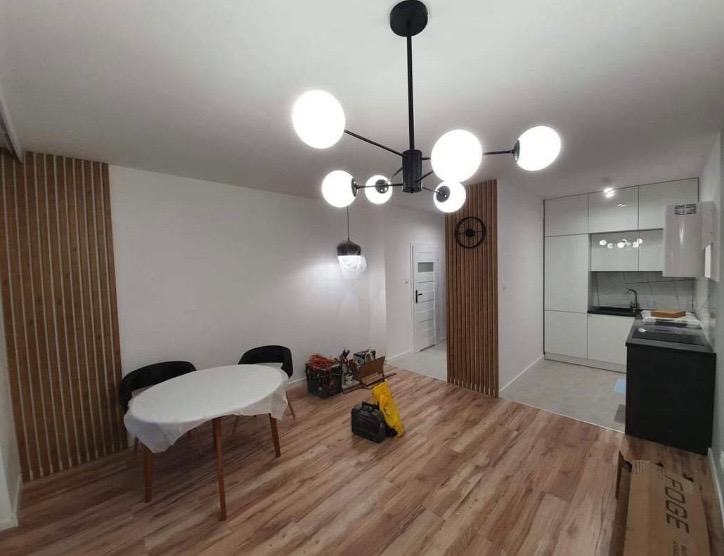 Jasne wnętrze po remoncie z drewnianą podłogą, panelami ściennymi i nowoczesną lampą sufitową z kulistymi kloszami, okrągły stół przykryty folią ochronną, widoczna kuchnia w tle