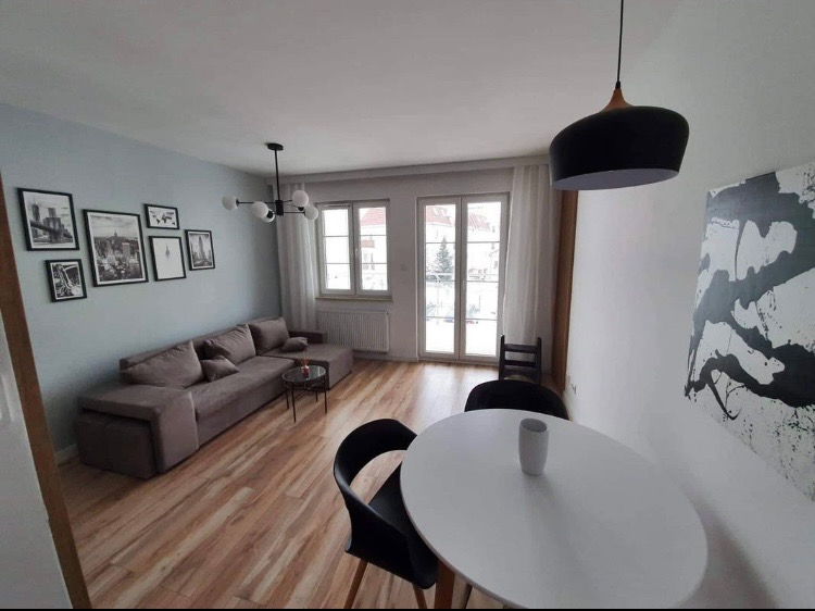 Jasne wnętrze salonu z szarą narożną sofą, okrągłym białym stołem, czarnymi krzesłami, drewnianą podłogą i galerią czarno-białych zdjęć na ścianie w kolorze gołębim.