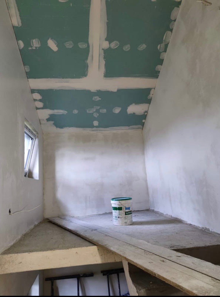 Pokój w trakcie wykańczania: sufit z płyt kartonowo-gipsowych z widocznymi łączeniami, szpachlowane ściany, wiadro z farbą na podłodze, drewniana kładka