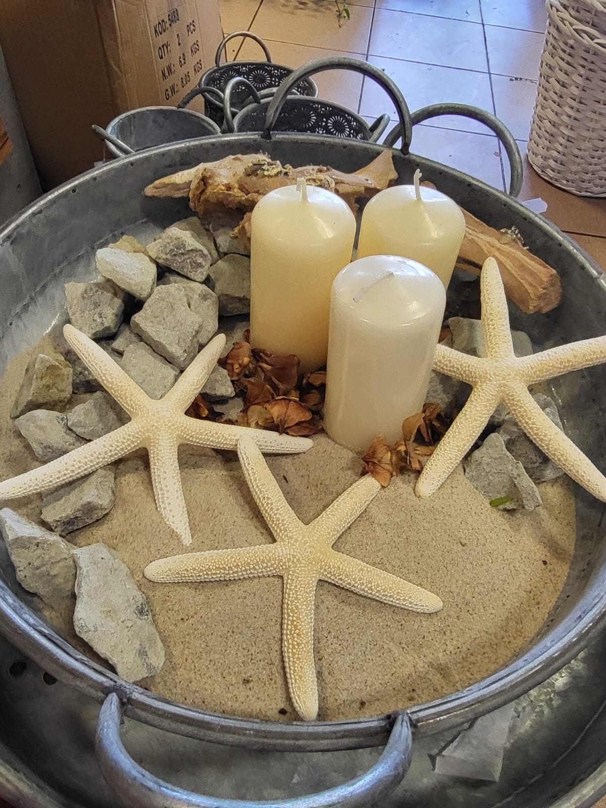 Dekoracja stołu w metalowej misie: trzy białe świece cylindryczne, trzy rozgwiazdy, kamienie, piasek i kawałki drewna.