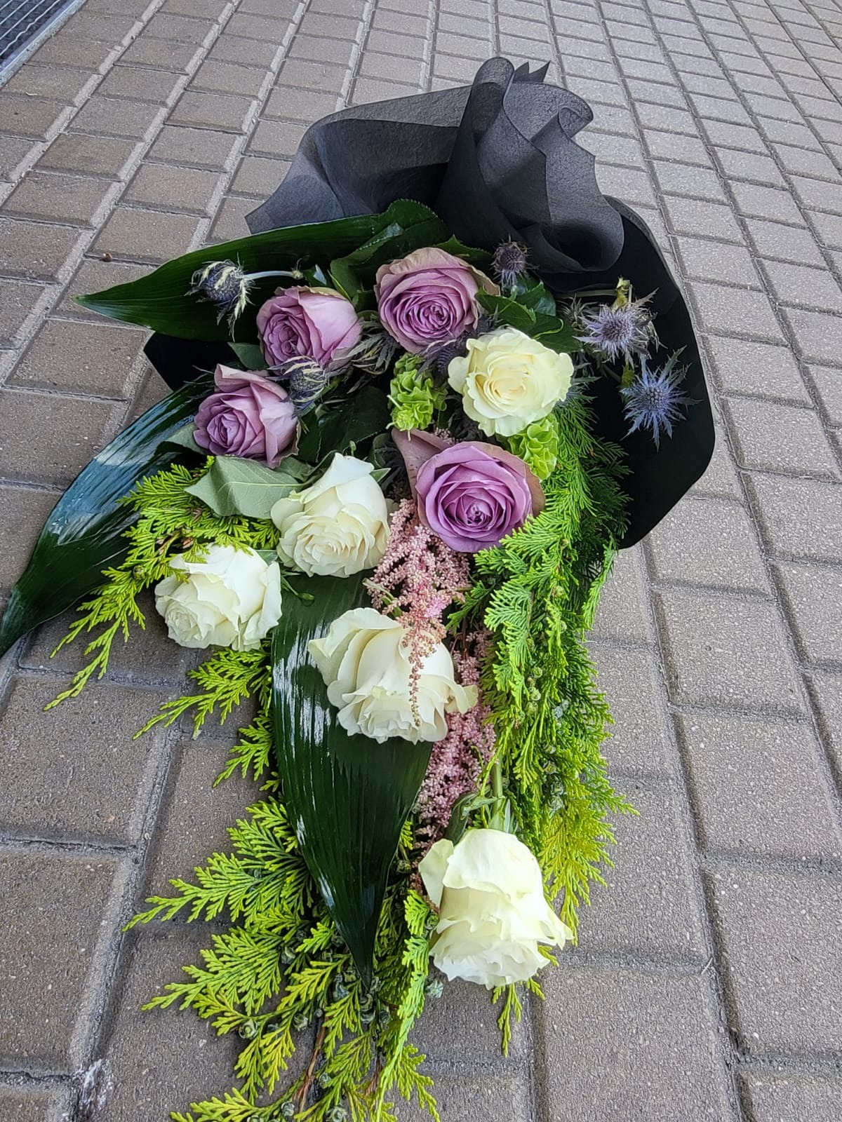 Kompozycja kwiatowa z białych i fioletowych róż, paproci i ostu, owinięta w czarny papier, leżąca na szarej kostce brukowej.