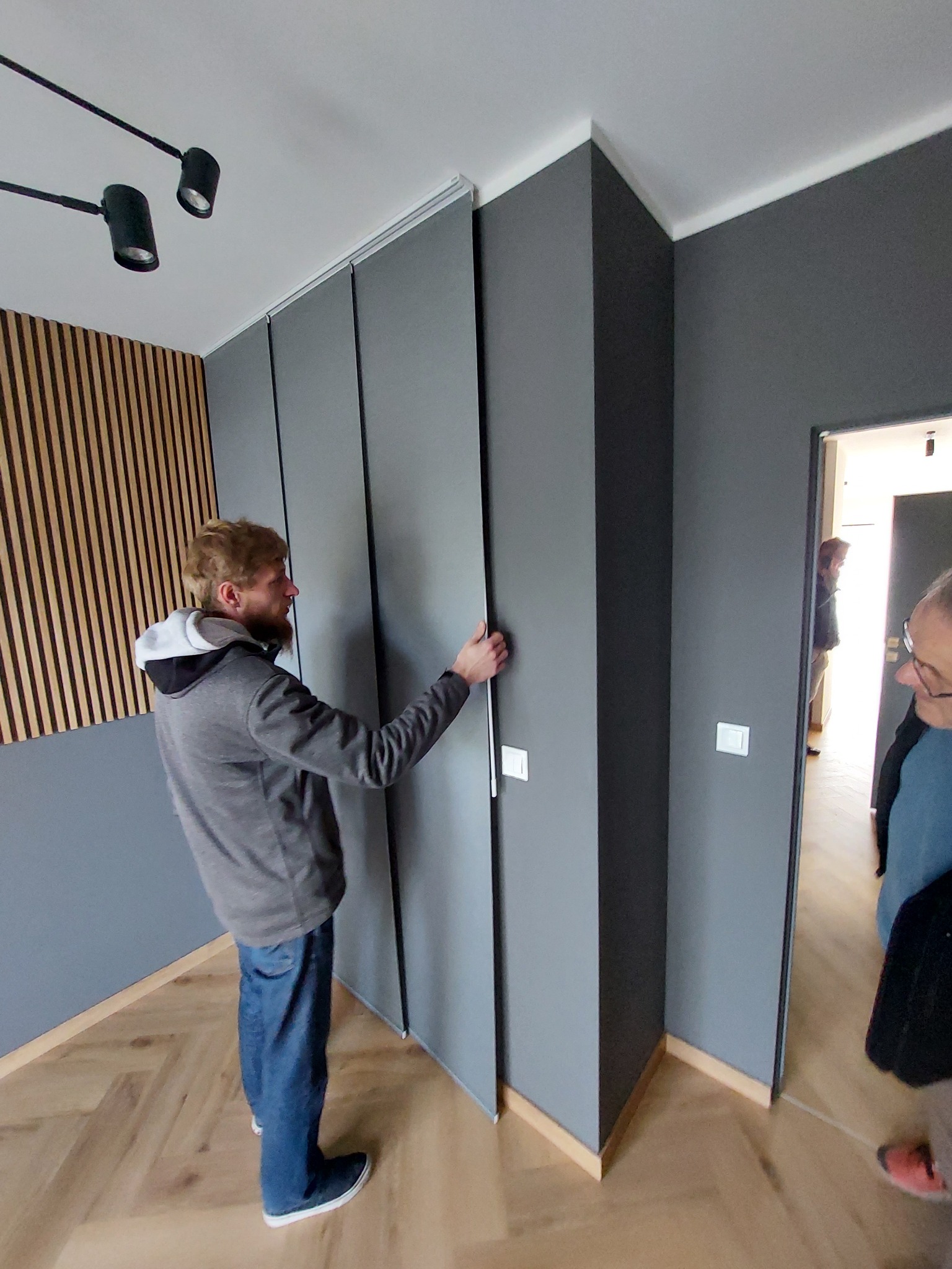 Mężczyzna w szarej bluzie przesuwa szare, przesuwne drzwi szafy wnękowej na tle ściany z drewnianymi lamelami i parkietem ułożonym w jodełkę.