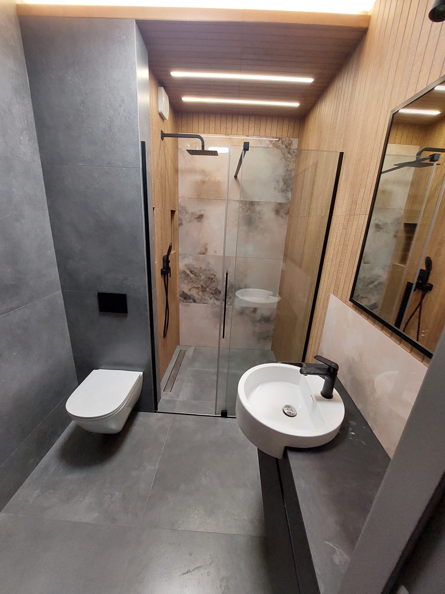 Nowoczesna łazienka z kabiną prysznicową typu walk-in, umywalką nablatową na ciemnym blacie, wiszącą toaletą i szarą płytką imitującą beton na ścianach oraz drewnianą okładziną na suficie...