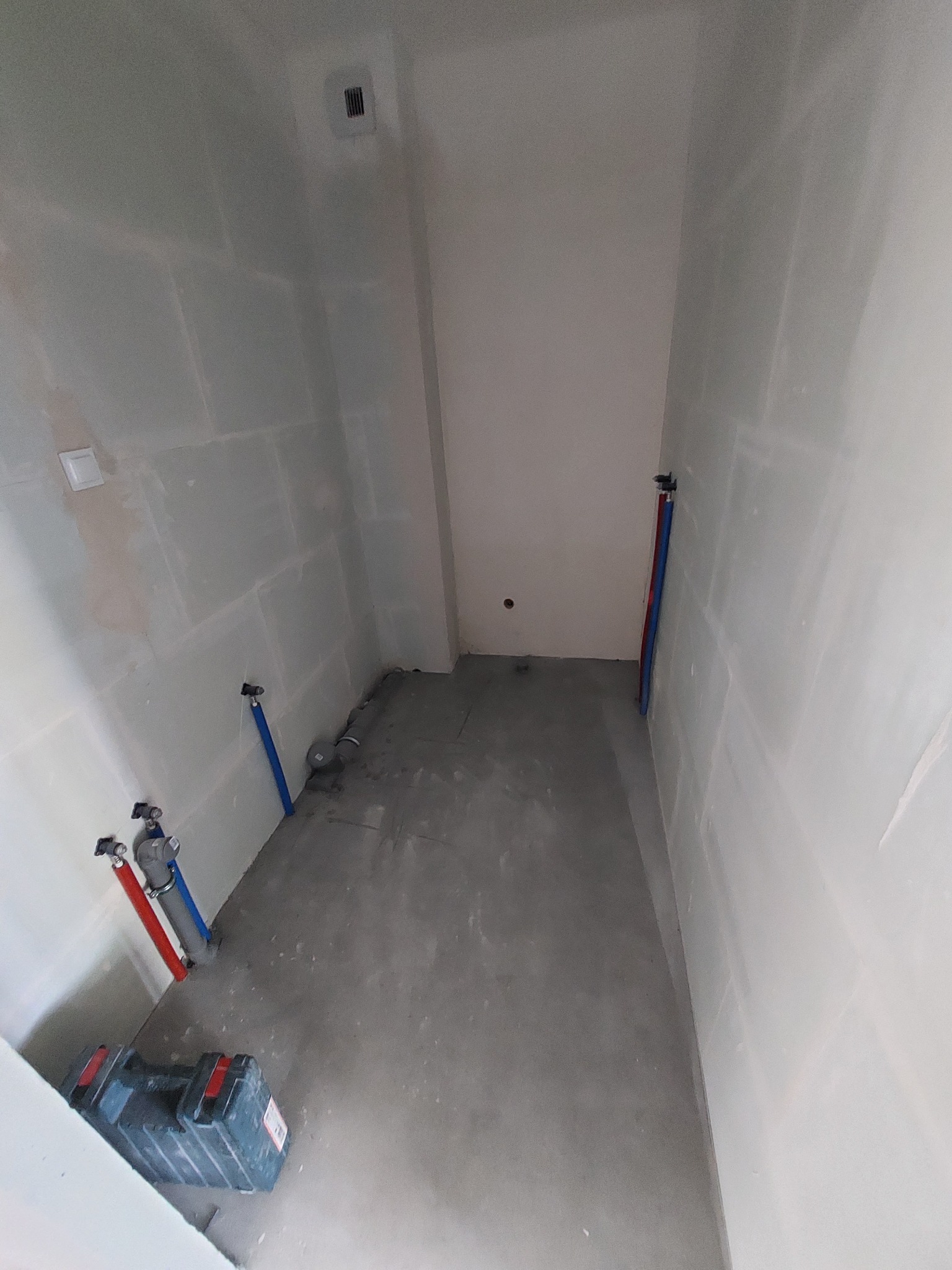 Wnętrze pomieszczenia w trakcie prac przygotowawczych: ściany wyłożone płytami gipsowo-kartonowymi, widoczne rury instalacji wodnej w kolorach czerwonym i niebieskim oraz skrzynka z narzędziami...