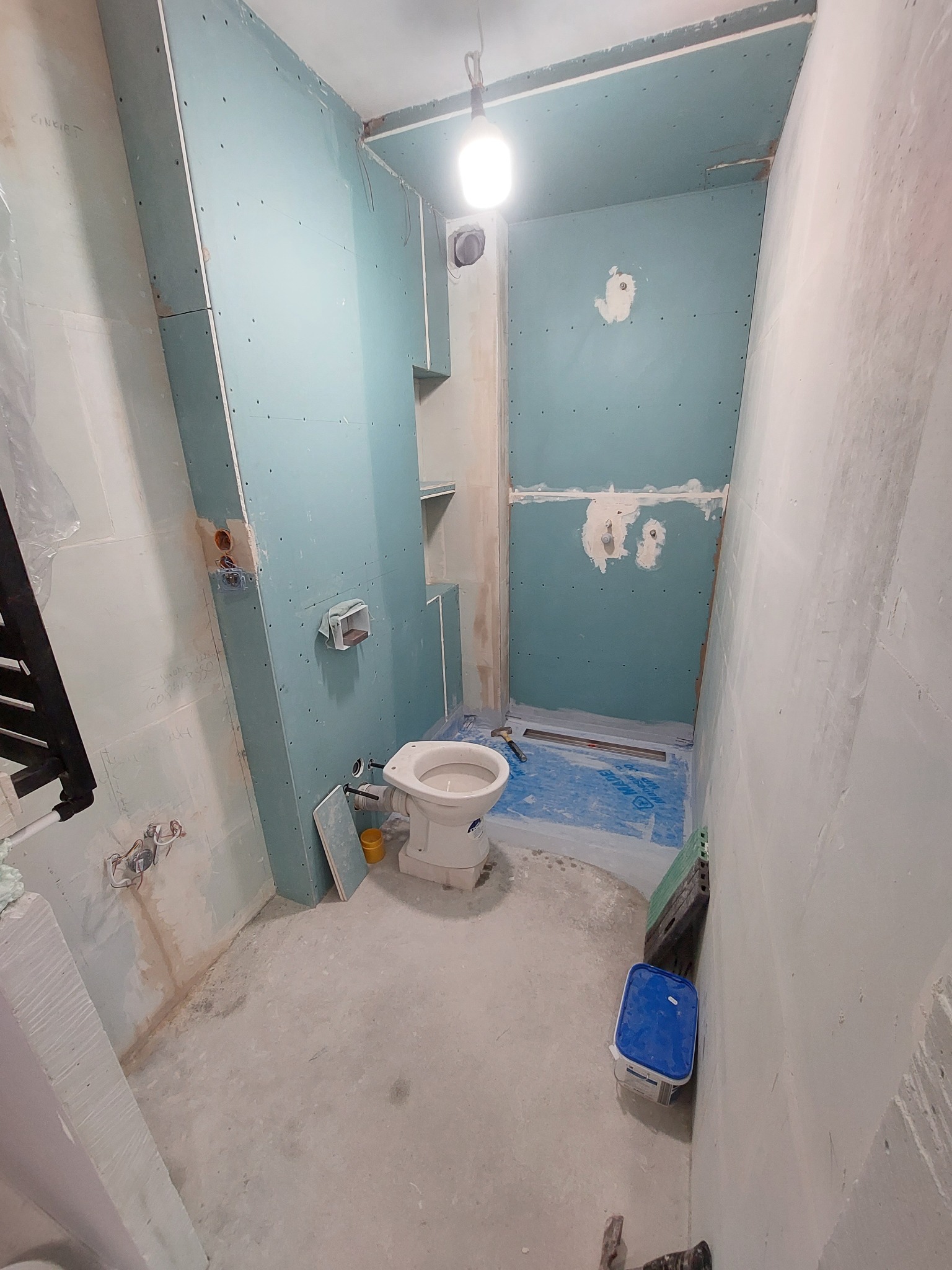 Łazienka w trakcie prac: montaż płyt gipsowo-kartonowych na ścianach, widoczny stelaż prysznica z odpływem liniowym, częściowo zamontowana toaleta, surowy beton na podłodze i ścianach, niebieskie...