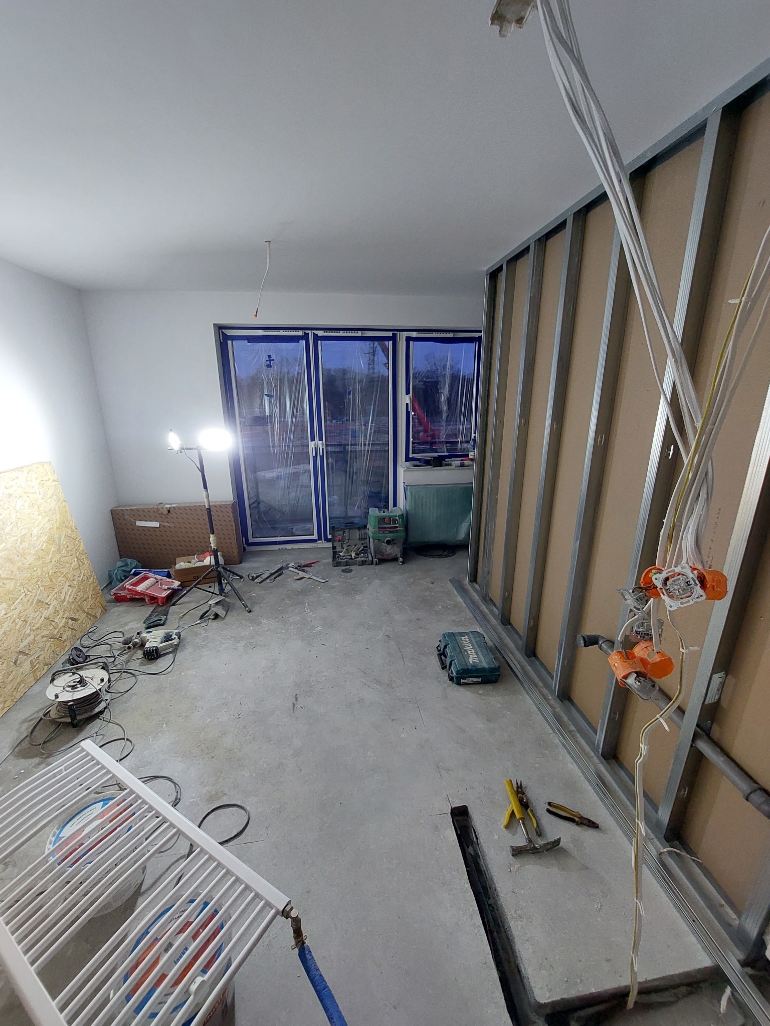 Wnętrze w trakcie prac wykończeniowych: widoczne profile ścian działowych, instalacja elektryczna w puszkach, grzejnik, narzędzia i materiały budowlane na surowej posadzce.
