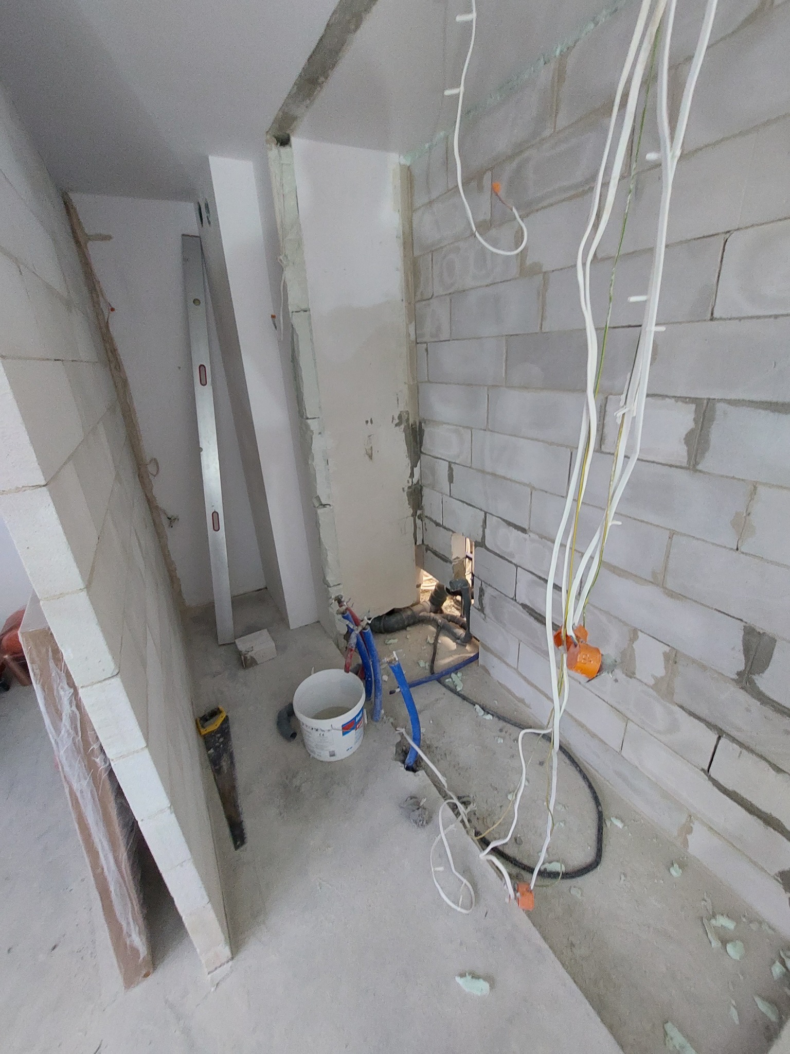 Wnętrze w trakcie remontu: ściana z białych bloczków, wystające kable elektryczne, fragment instalacji hydraulicznej z niebieskimi rurami, poziomica oparta o ścianę, wiadro z zaprawą i piła ręczna...