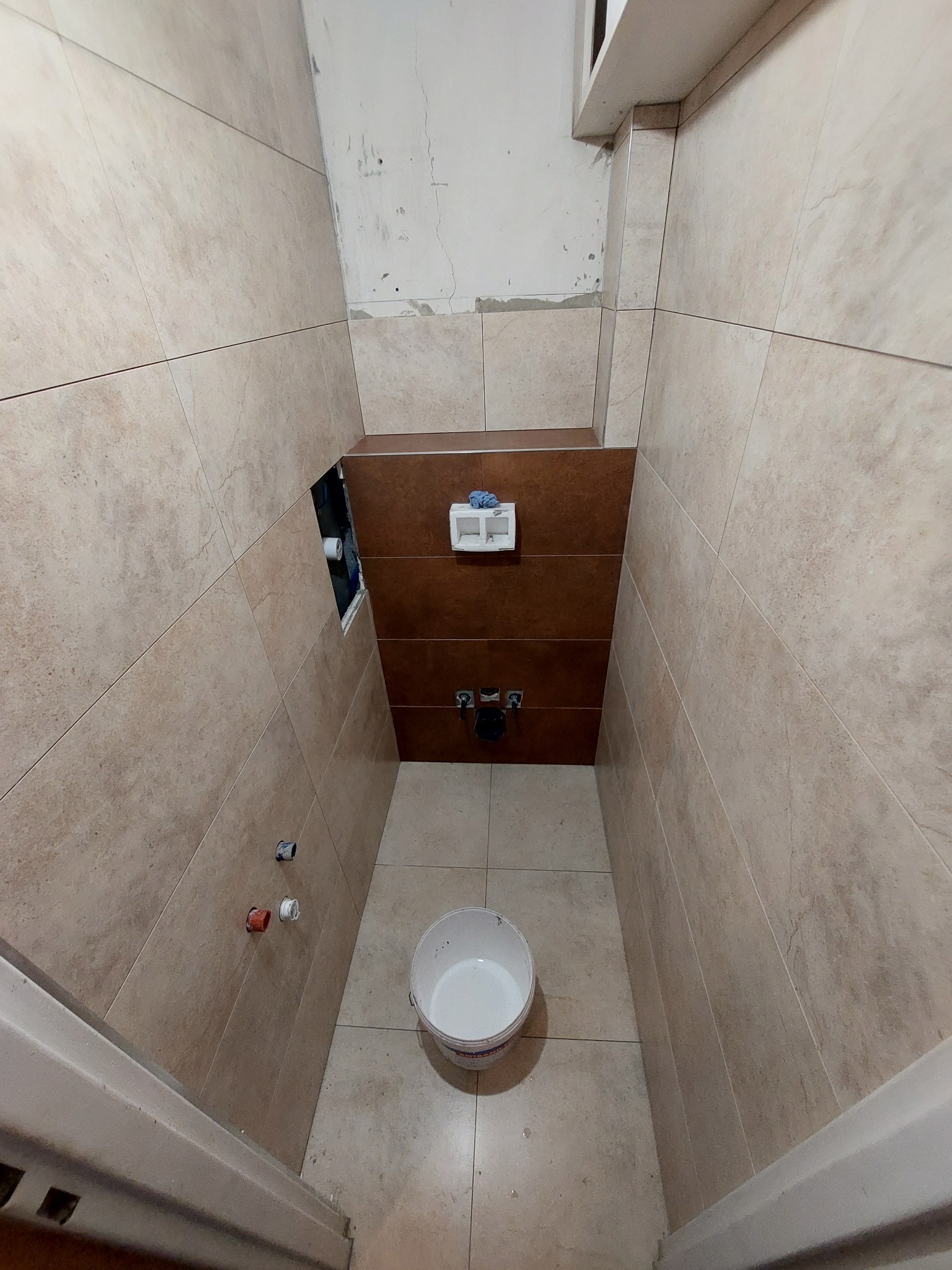 Wąska łazienka w trakcie kładzenia beżowych płytek ściennych i podłogowych, z fragmentem brązowych płytek nad instalacją wodną, widoczny otwór w ścianie, białe wiadro na podłodze, surowy stan...