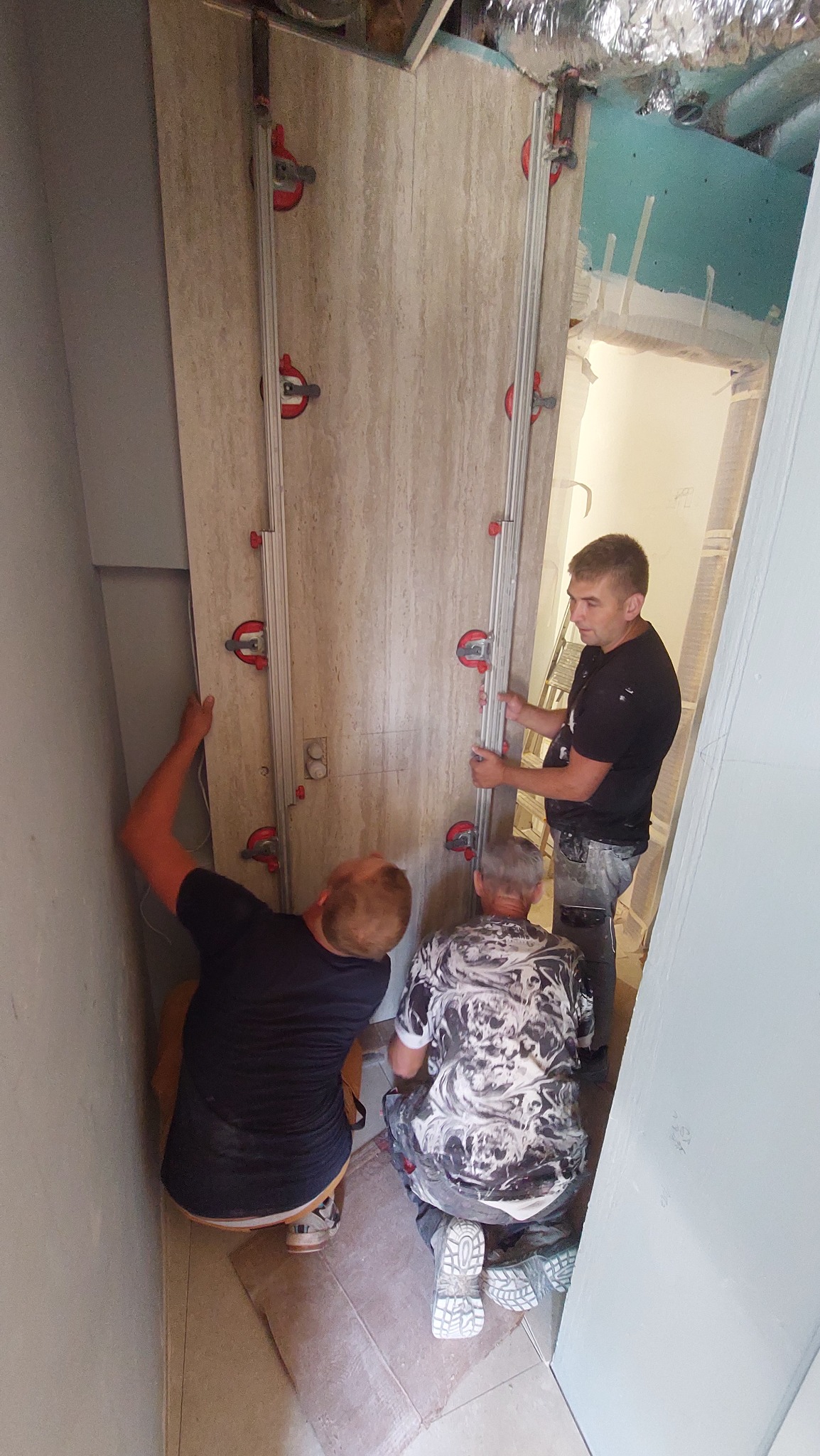 Trzech mężczyzn montuje dużą płytę z imitacją trawertynu na ścianie, używając przyssawek z czerwoną dźwignią i prowadnic. W tle widoczna instalacja wentylacyjna i surowa ściana z płyt...