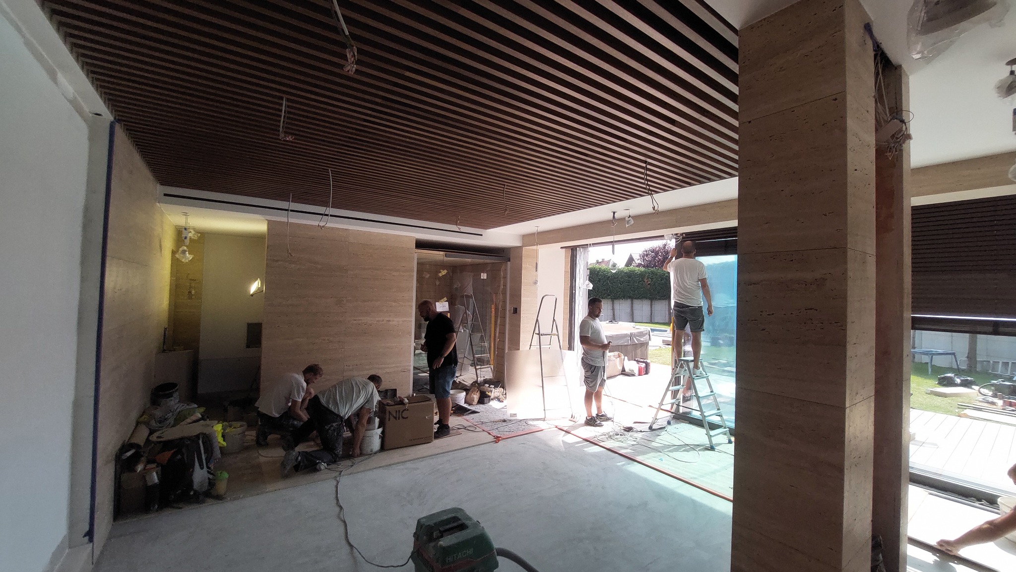 Wnętrze w trakcie prac wykończeniowych: montaż drewnianego sufitu lamelowego, ściany wyłożone trawertynem, ekipa remontowa w trakcie pracy, widoczny odkurzacz przemysłowy i drabiny.