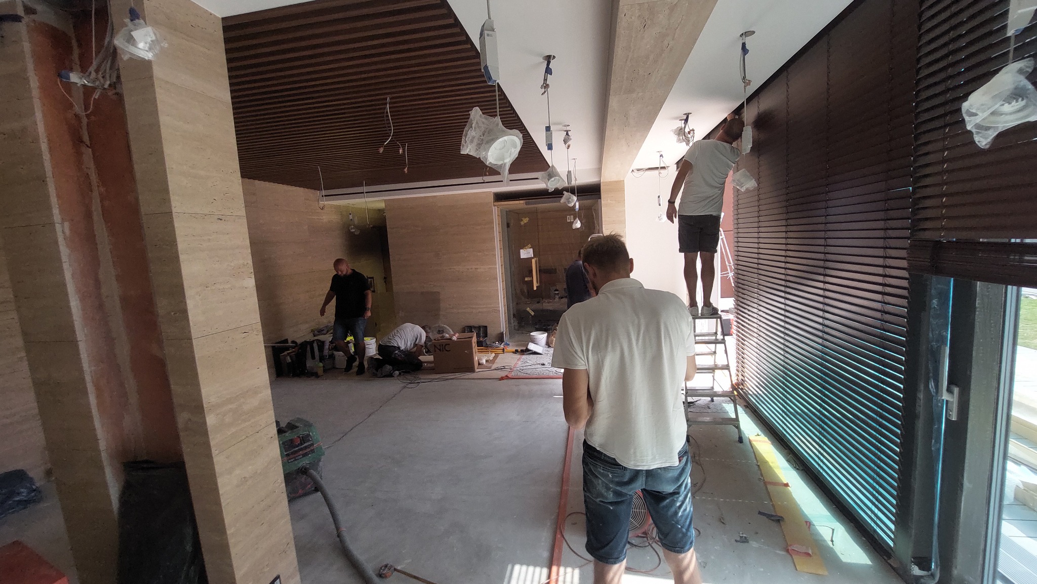 Montaż drewnianych żaluzji w nowoczesnym wnętrzu z marmurowymi kolumnami, widoczne okablowanie i ekipa remontowa w trakcie prac wykończeniowych.