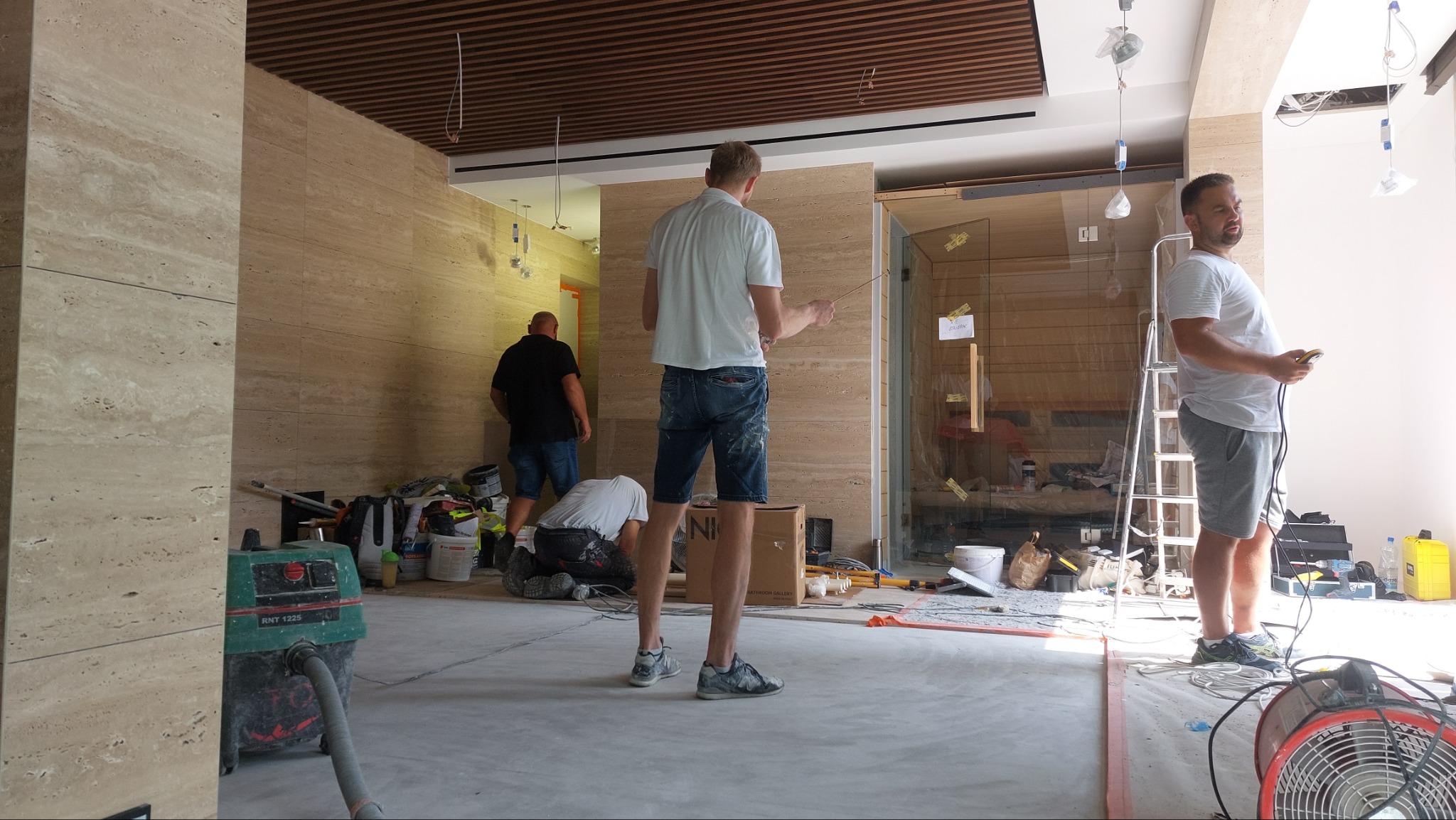 Wnętrze w trakcie prac wykończeniowych: ściany wyłożone trawertynem, widoczni trzej pracownicy, jeden z nich mierzy ścianę, drugi pracuje na podłodze, trzeci stoi przy szklanej kabinie, na podłodze...