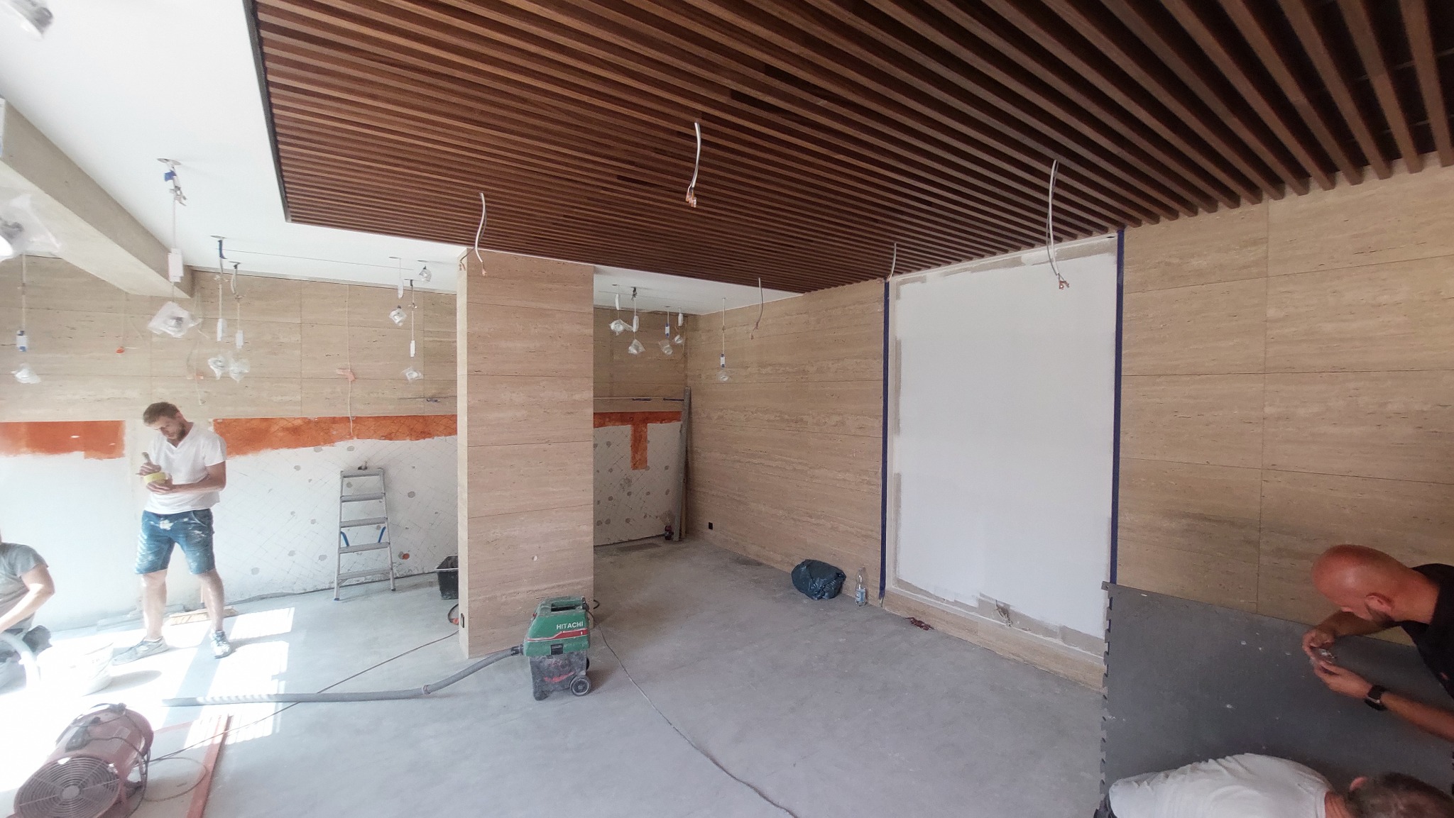 Montaż drewnianego sufitu lamelowego w pomieszczeniu z wykończonymi ścianami z trawertynu, widoczni dwaj pracownicy podczas prac wykończeniowych, jeden z nich miesza zaprawę, drugi zajmuje się...