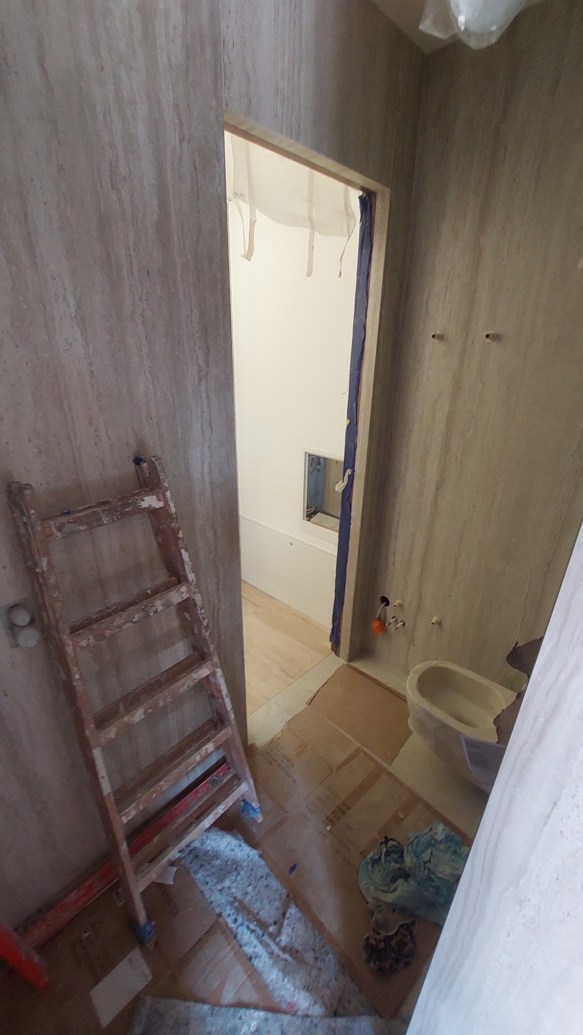 Łazienka w trakcie remontu: widoczna drabina oparta o ścianę z imitacji trawertynu, zabezpieczona podłoga kartonem, częściowo zamontowana toaleta i widok na wannę w sąsiednim pomieszczeniu.