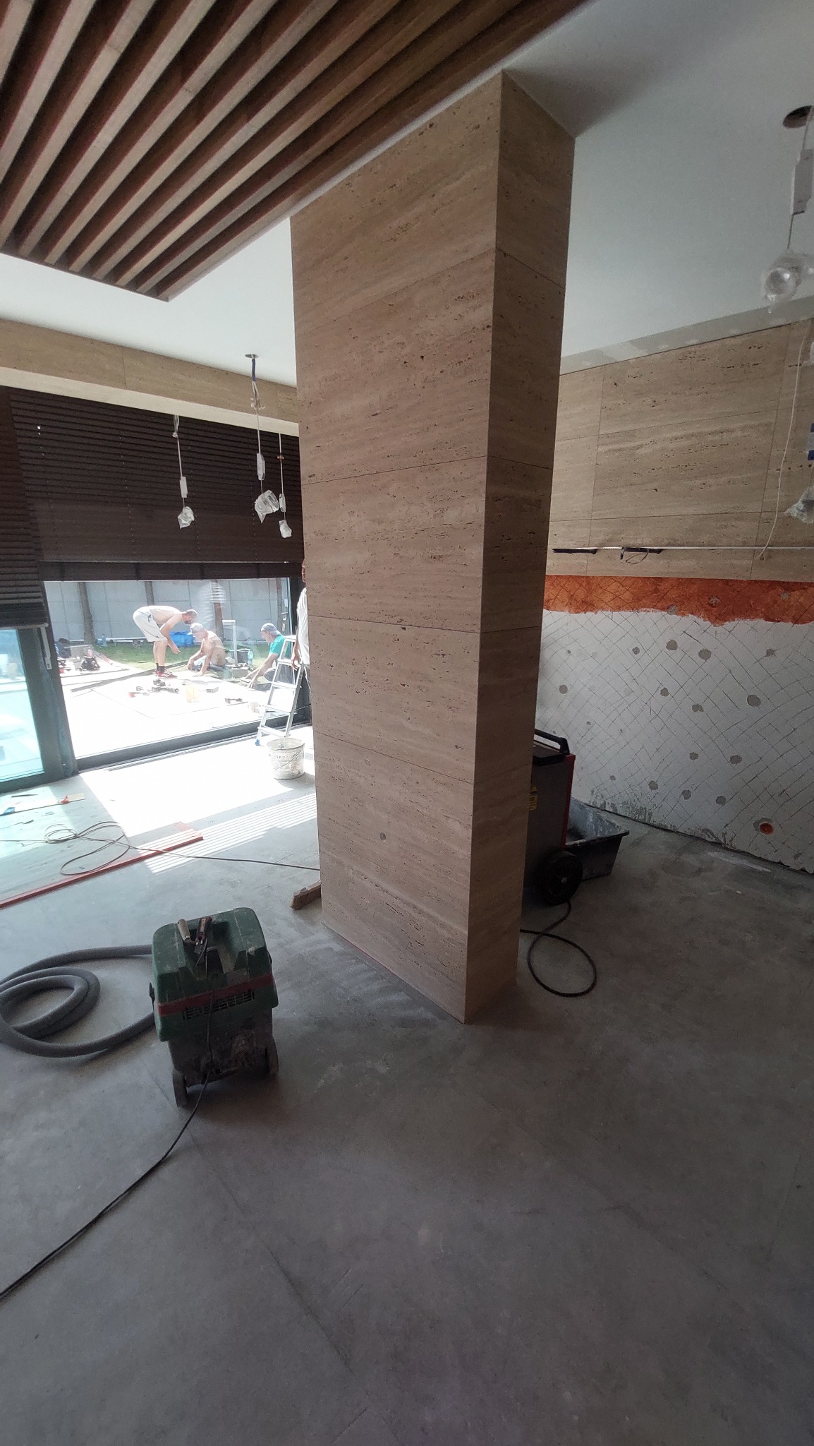 Montaż kolumny z płytek imitujących trawertyn w surowym wnętrzu, widoczny odkurzacz przemysłowy, w tle okno z widokiem na prace w ogrodzie.