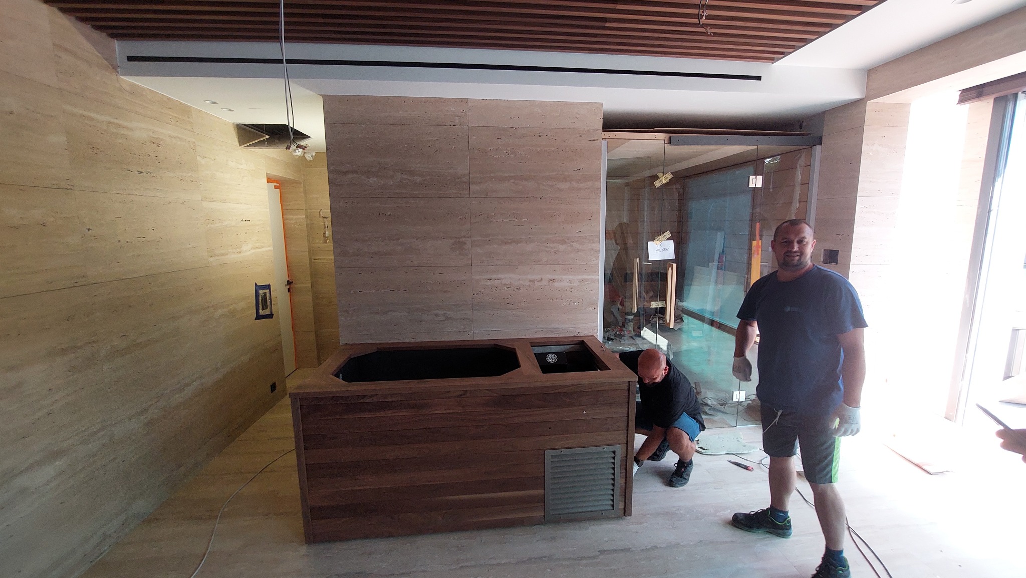 Montaż drewnianej obudowy wanny z hydromasażem w łazience wykończonej trawertynem, widoczni dwaj pracownicy w trakcie instalacji, jeden z nich uśmiecha się do kamery.