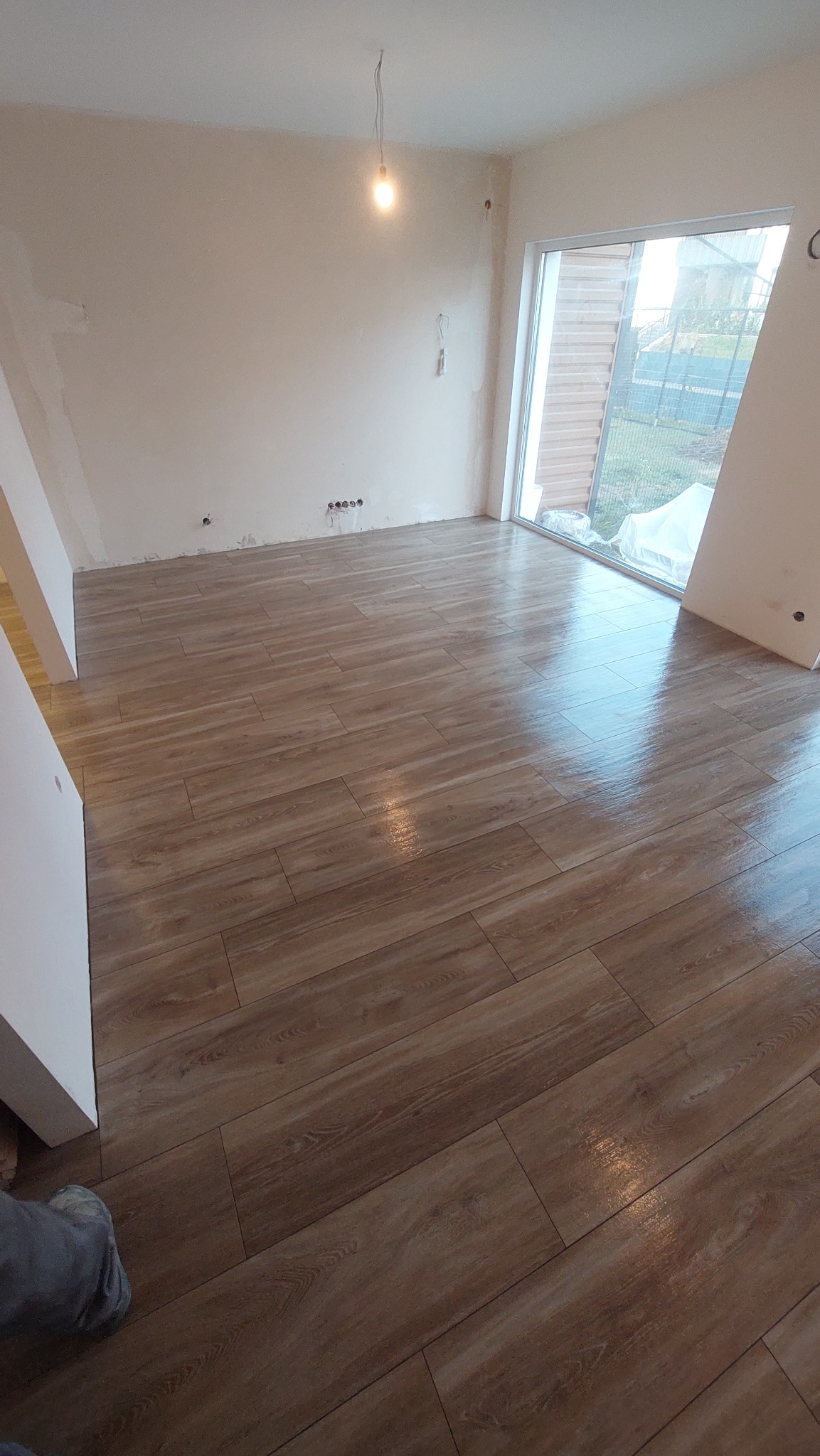 Ułożona podłoga z paneli imitujących drewno w surowym pomieszczeniu, widoczne ślady po gładzi na ścianach, okno z widokiem na zewnątrz, na podłodze delikatne refleksy świetlne.