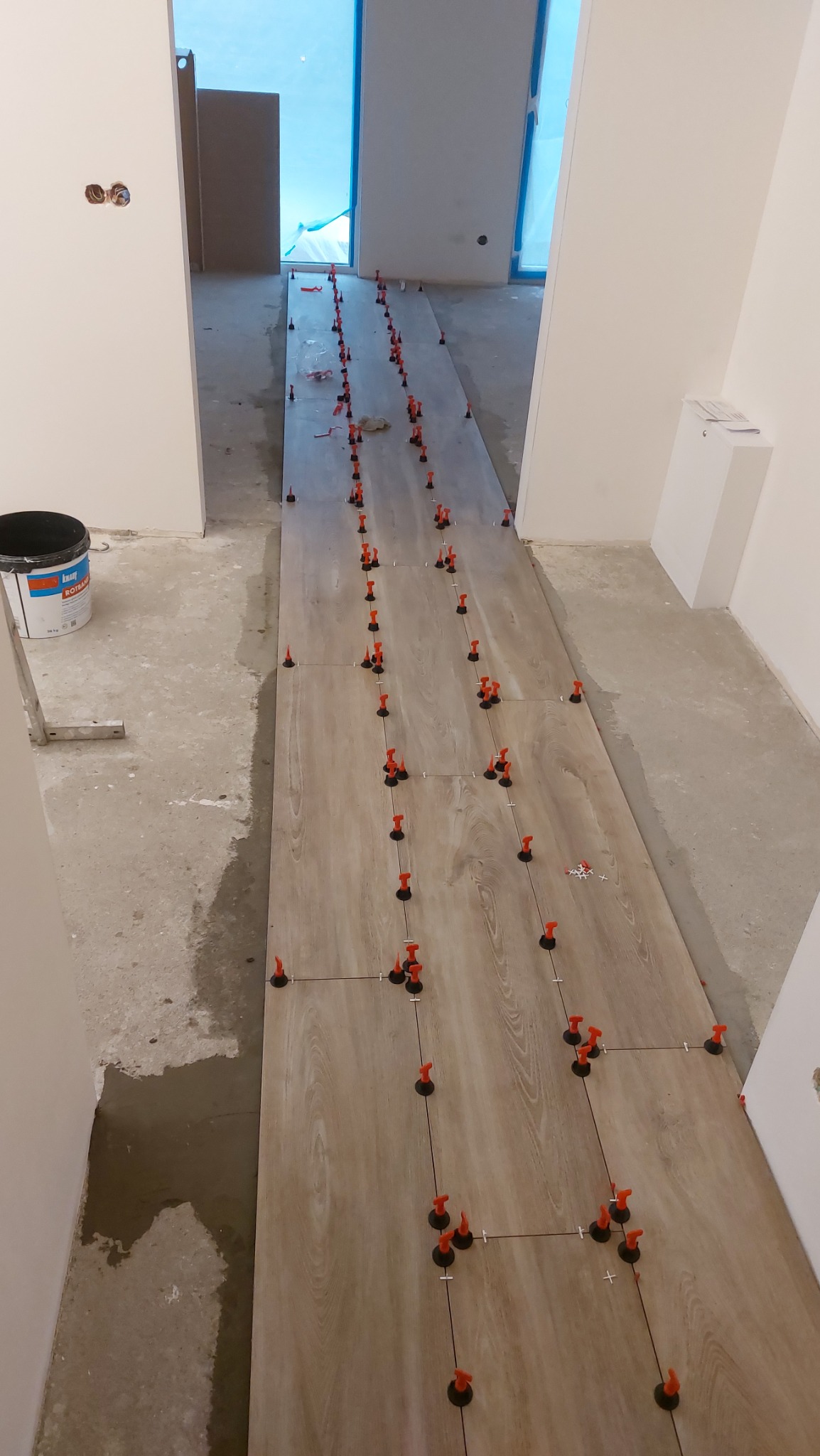 Układanie płytek podłogowych imitujących drewno w korytarzu, z użyciem systemu poziomowania z czerwonymi klipsami, widoczny kubeł z klejem na podłodze.