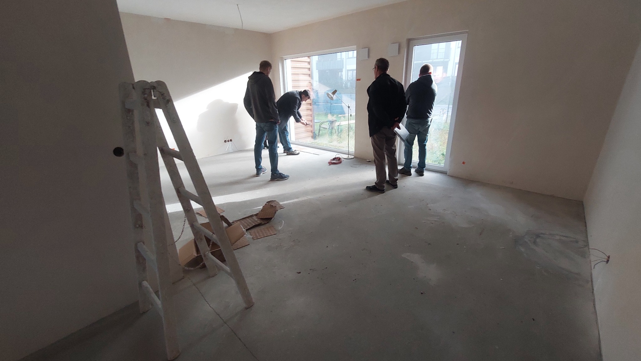 Wnętrze w stanie deweloperskim z grupą mężczyzn, jeden pracuje przy oknie, widoczna drabina malarska, surowy beton na podłodze, ściany przygotowane do malowania.