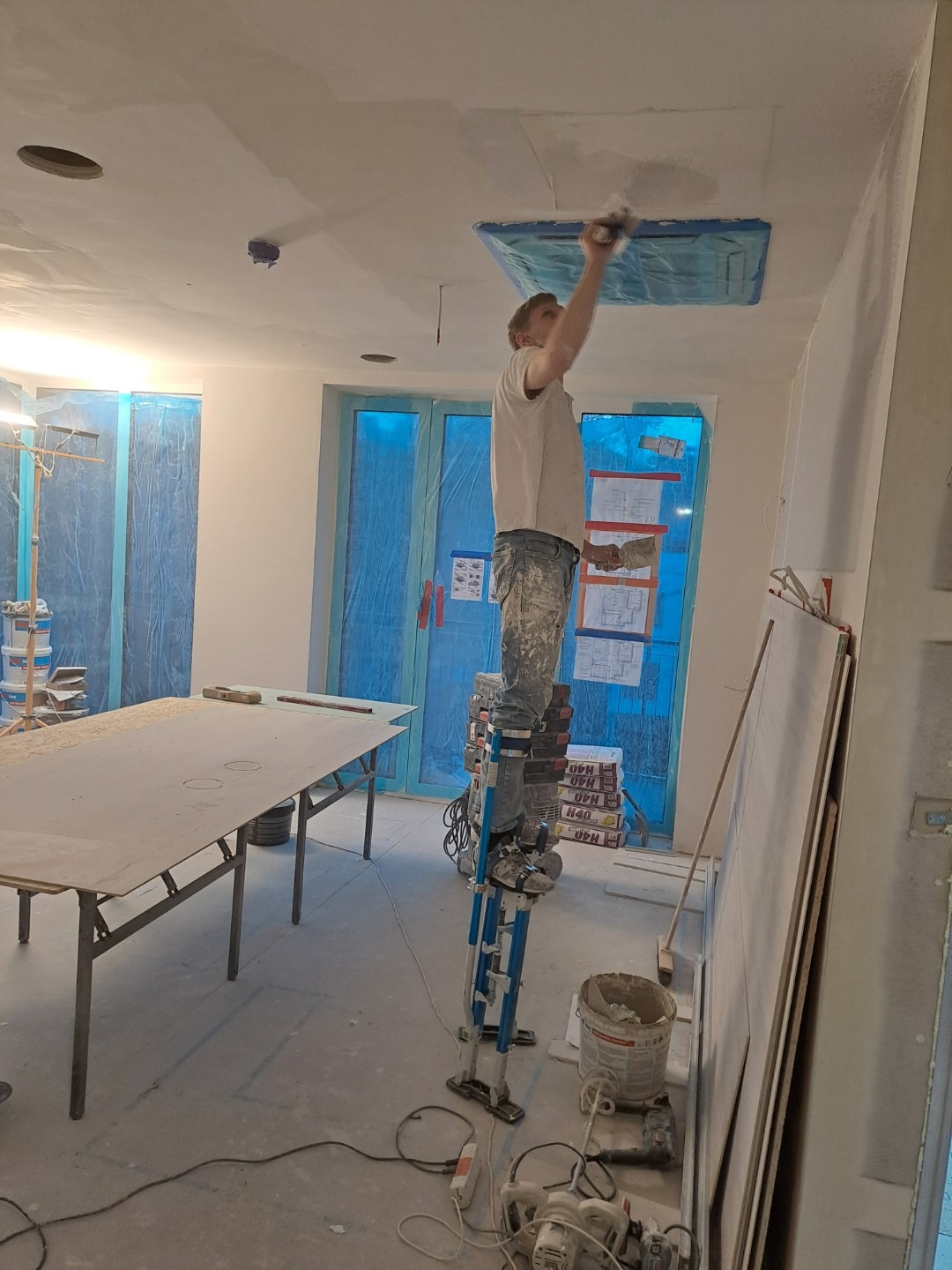 Pracownik na szczudłach wykończeniowych szpachluje sufit w pomieszczeniu z zabezpieczonymi folią oknami, widoczne narzędzia i materiały budowlane.
