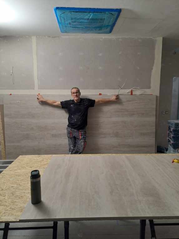 Mężczyzna w czarnej koszulce i zabrudzonych spodniach, z rozłożonymi ramionami, prezentuje montaż paneli ściennych o fakturze jasnego marmuru, widoczne narzędzia i zabezpieczona folią klimatyzacja...