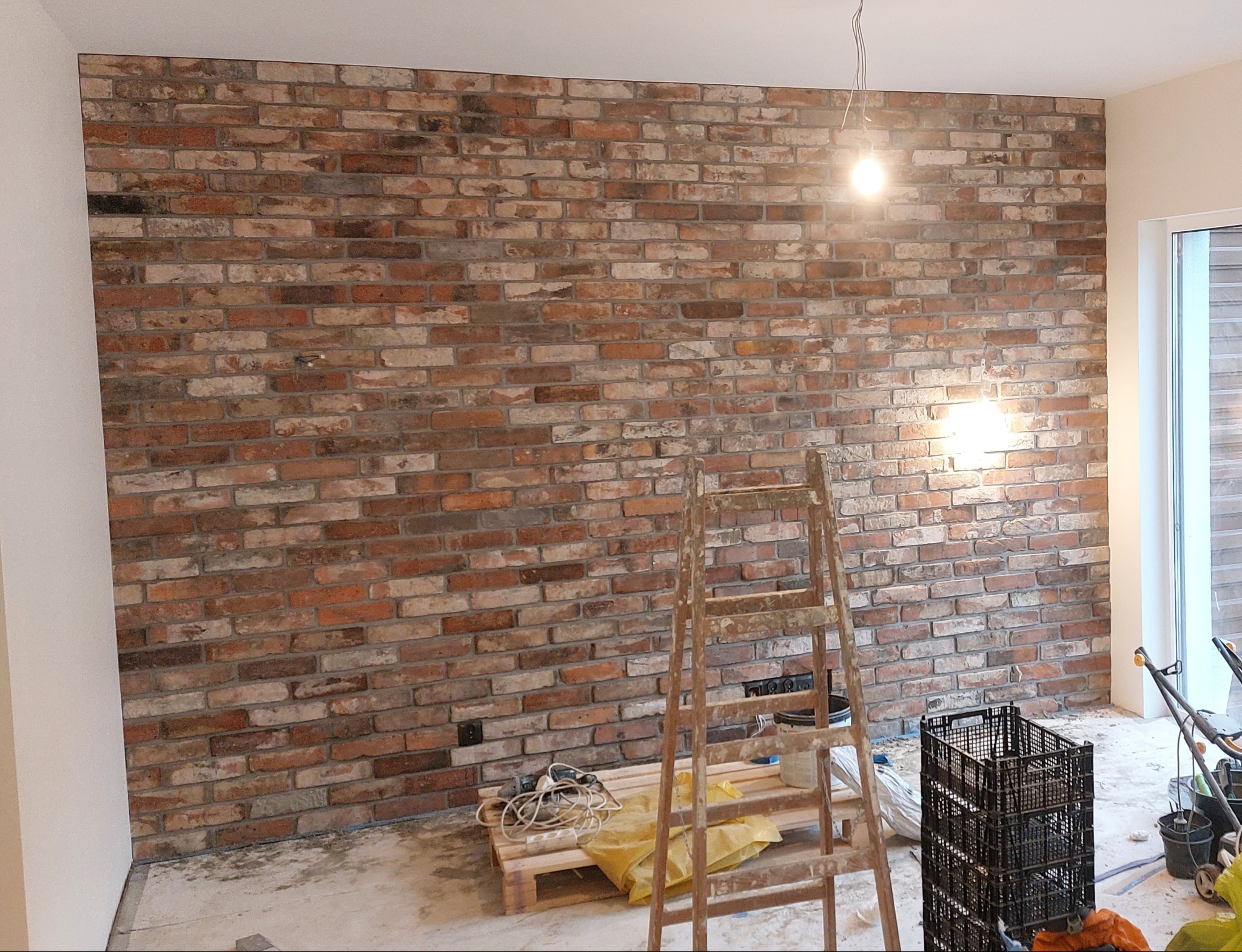 Ściana z cegły klinkierowej w trakcie montażu, widoczne narzędzia, drabina i nieporządek na podłodze.