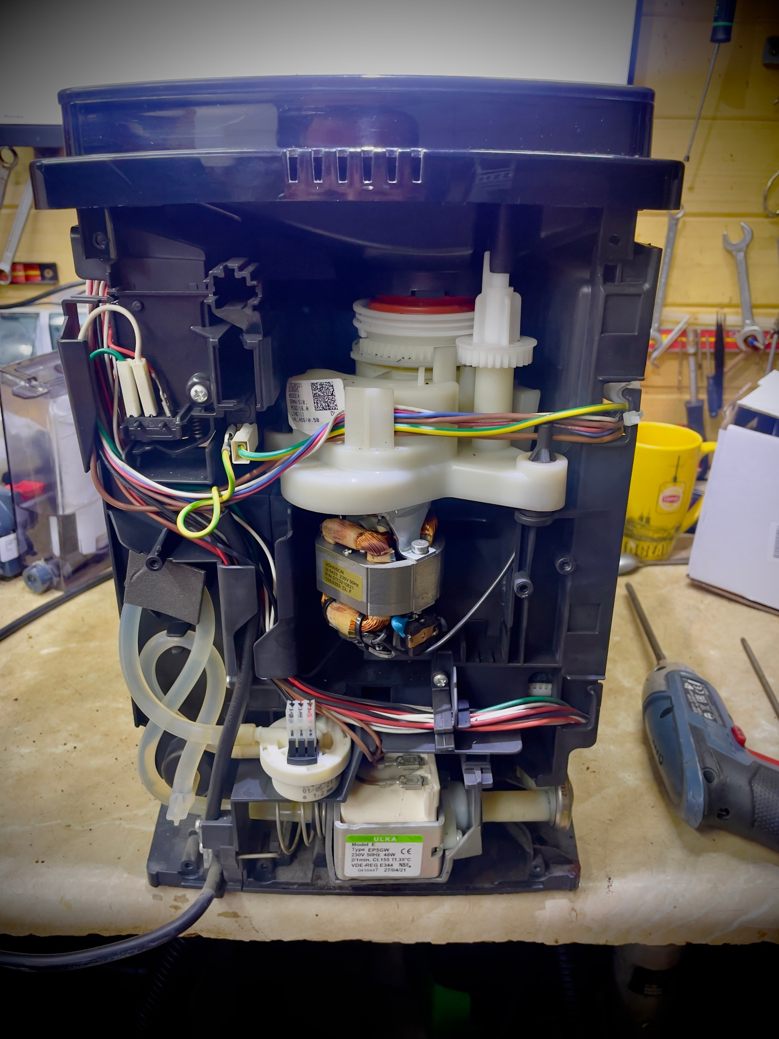 Wnętrze rozmontowanego ekspresu domowego z widocznymi elementami mechanicznymi, przewodami i silnikiem, na tle warsztatu z narzędziami.