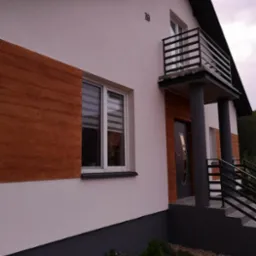 Biała elewacja domu z drewnopodobną okładziną, szarym cokołem i balkonem z czarną balustradą, numer domu '19' nad wejściem.