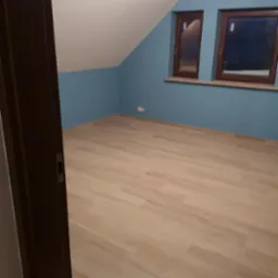 Poddasze po remoncie: pomalowane na niebiesko ściany, drewniane okna, jasna podłoga z paneli, widok zza ciemnych drzwi.