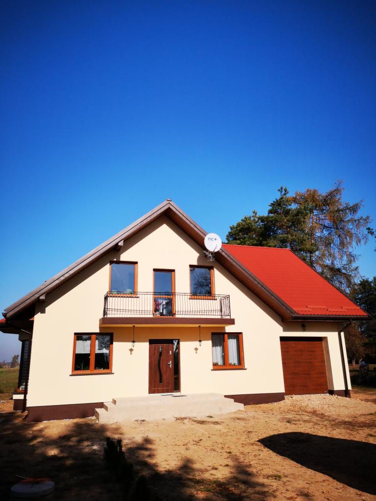 Nowo wybudowany dom jednorodzinny z beżową elewacją, czerwonym dachem i brązową stolarką okienną, widziany w słoneczny dzień z perspektywy niskiego kąta.