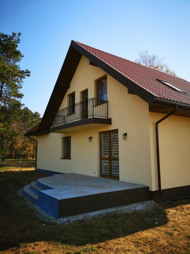 Żółty dom jednorodzinny z ciemnobrązową podmurówką i dachem, z balkonem z czarną balustradą, widok z zewnątrz w słoneczny dzień.