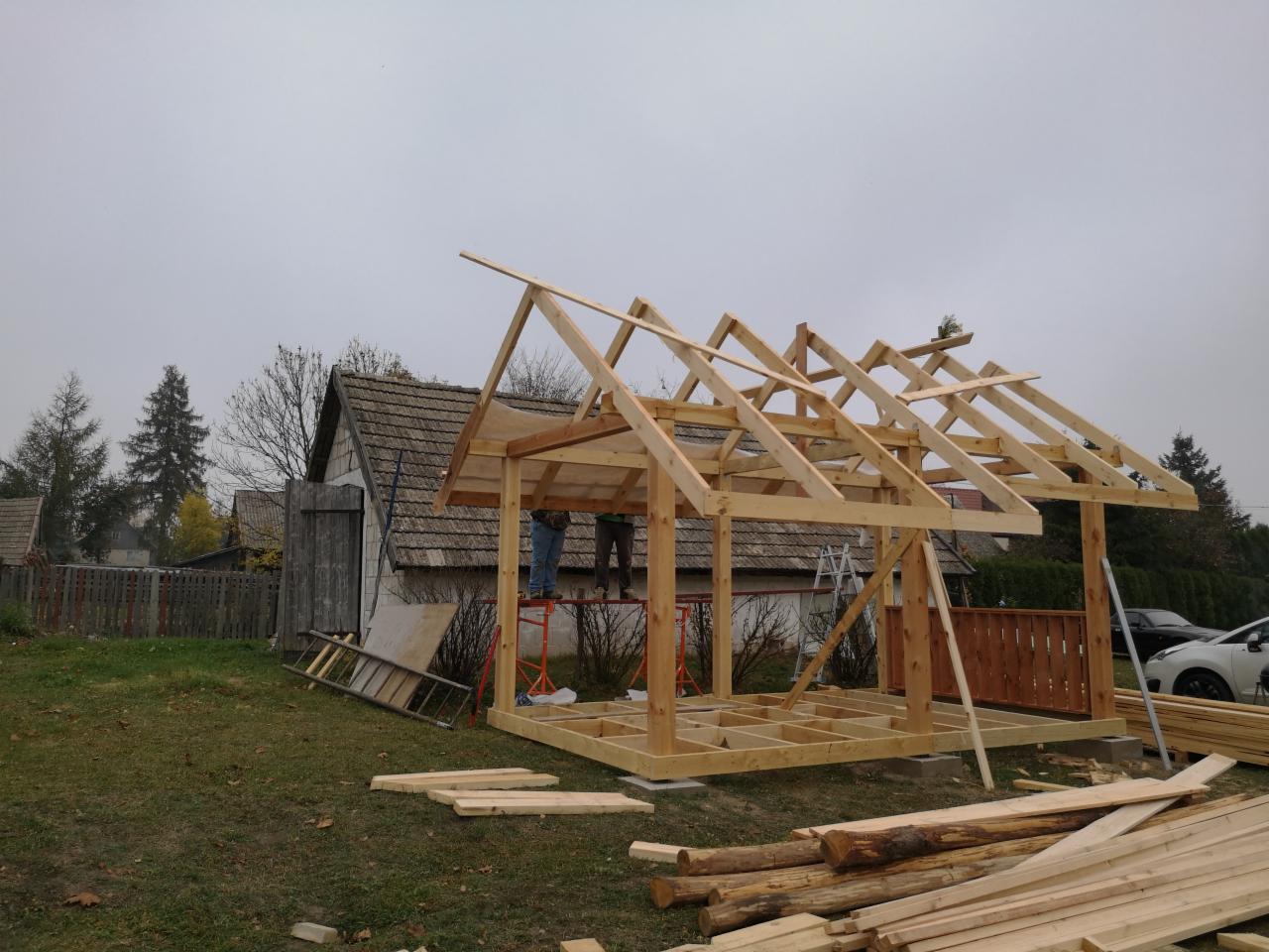 Budowa drewnianej altany ogrodowej w trakcie realizacji, widoczny szkielet konstrukcji dachu i podłogi, na tle starego domu i pochmurnego nieba.