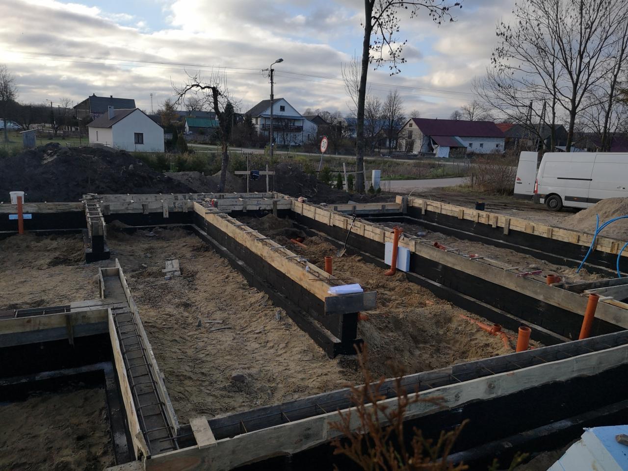 Przygotowanie fundamentów pod dom jednorodzinny: szalunki zalane piaskiem, izolacja pionowa, wystające rury kanalizacyjne, widok na zabudowę jednorodzinną w tle.