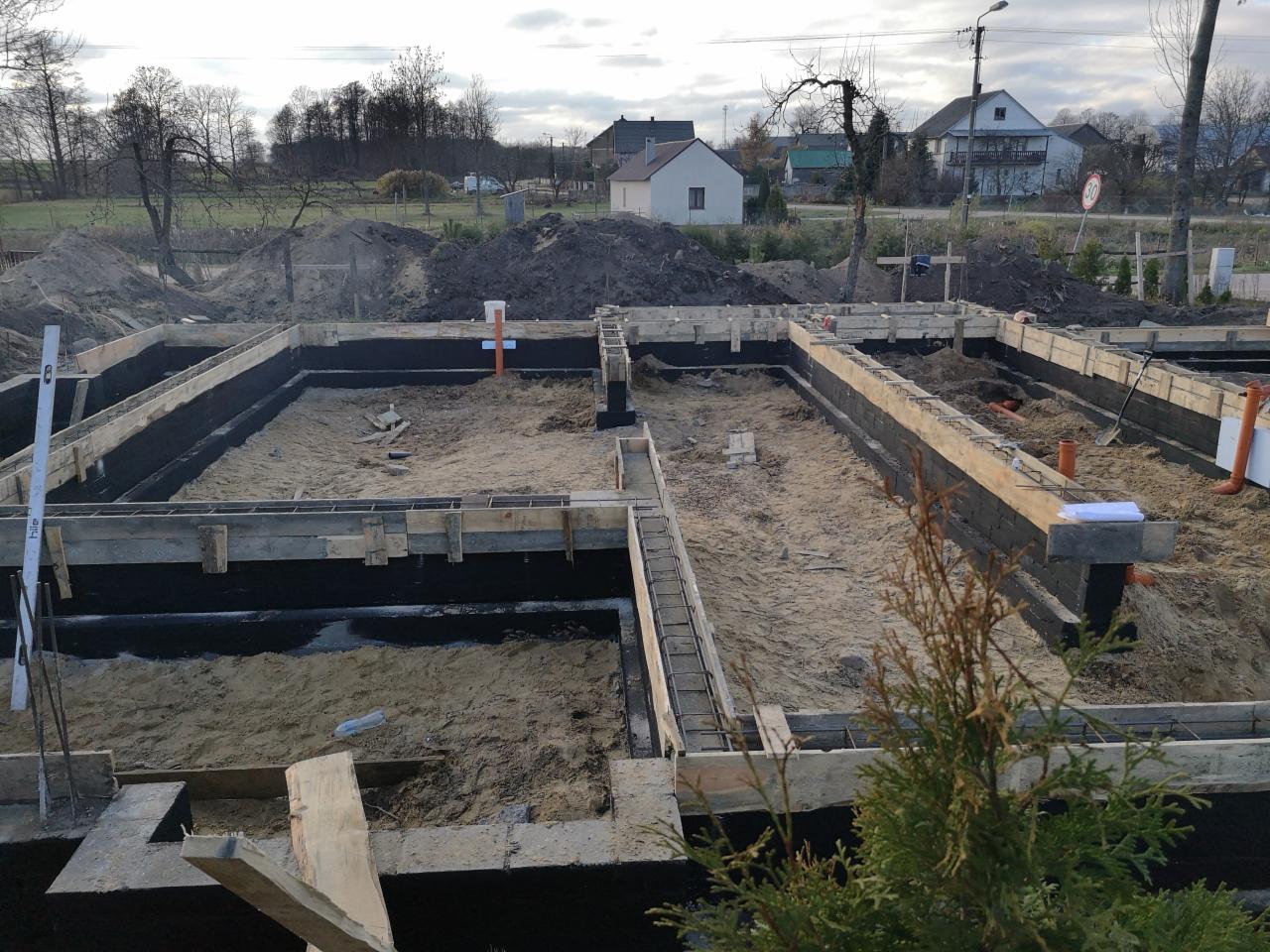Fundamenty domu w trakcie budowy: szalunki z drewna, izolacja przeciwwilgociowa, piasek zasypowy, widoczne rury instalacyjne, w tle zabudowa jednorodzinna.