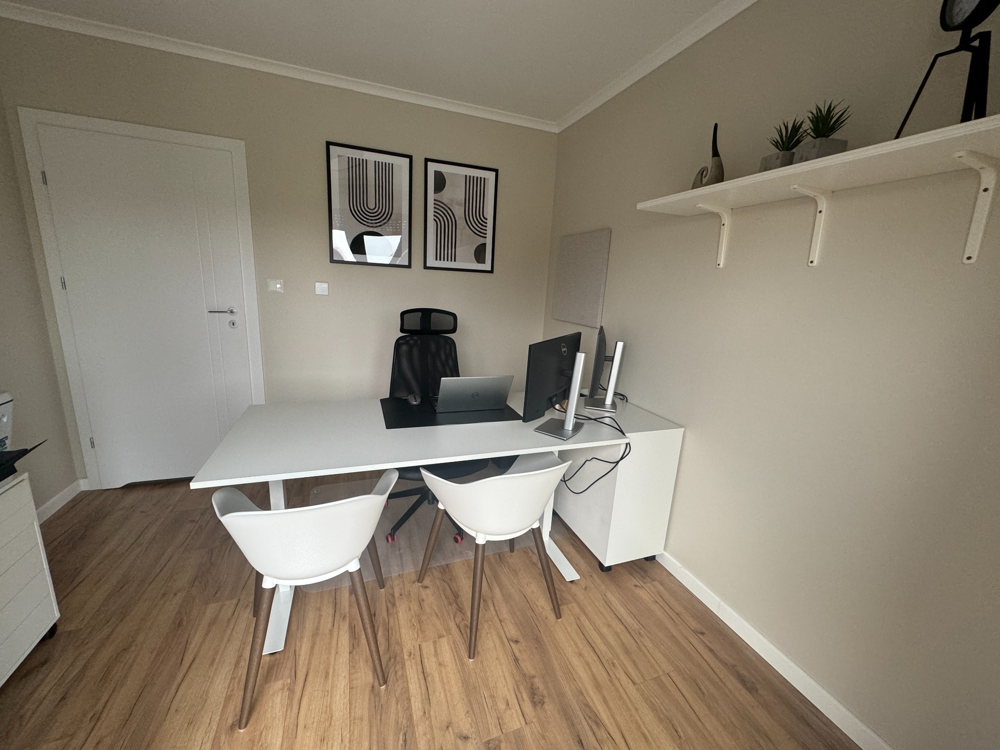 Nowoczesne biuro z białym biurkiem, krzesłami i laptopem, minimalistyczne dekoracje ścienne i półka z roślinami.