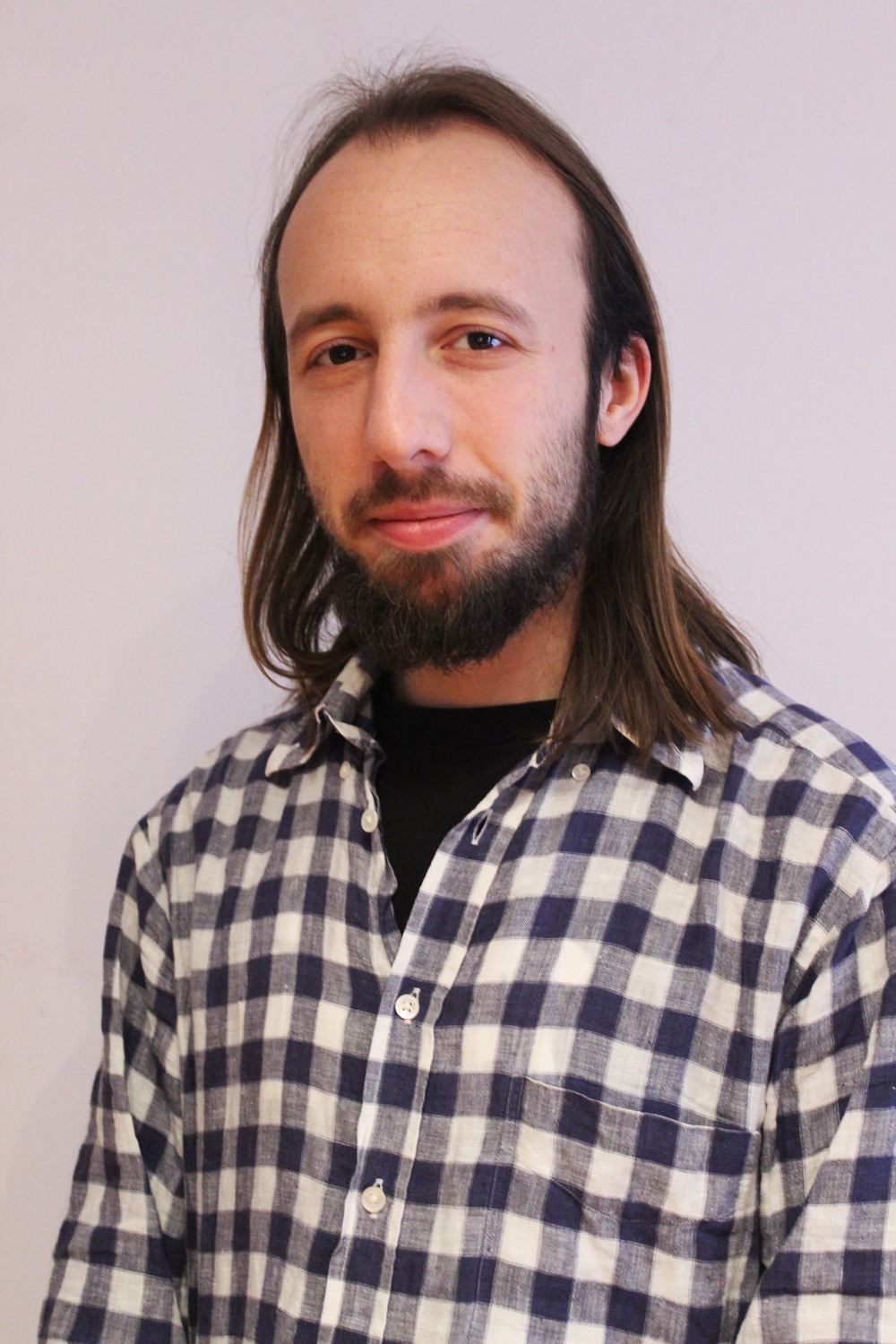 Portret mężczyzny z brodą i długimi włosami, ubranego w koszulę w kratę na tle jasnej ściany.