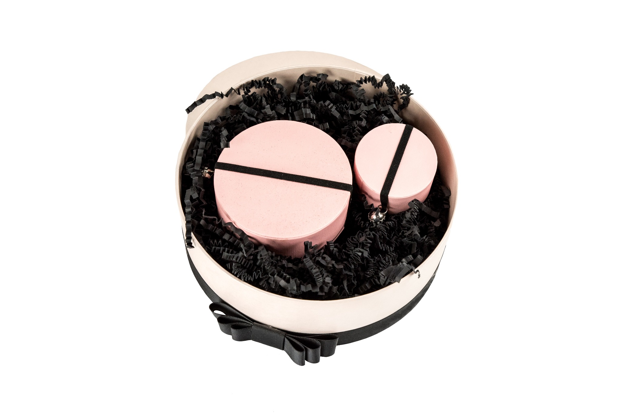 Otwarta, okrągła, kremowa pudełko z czarną kokardą, wypełniona czarnym papierem strzępkowym, zawierająca dwa różowe, okrągłe pudełka z czarnymi gumkami