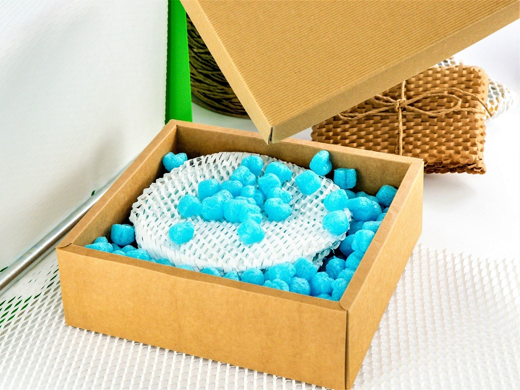 Otwarta, kwadratowa, kartonowa skrzynka wypełniona niebieskimi piankowymi wypełniaczami i białą siatką ochronną, z widoczną pokrywą i innymi materiałami zabezpieczającymi w tle.