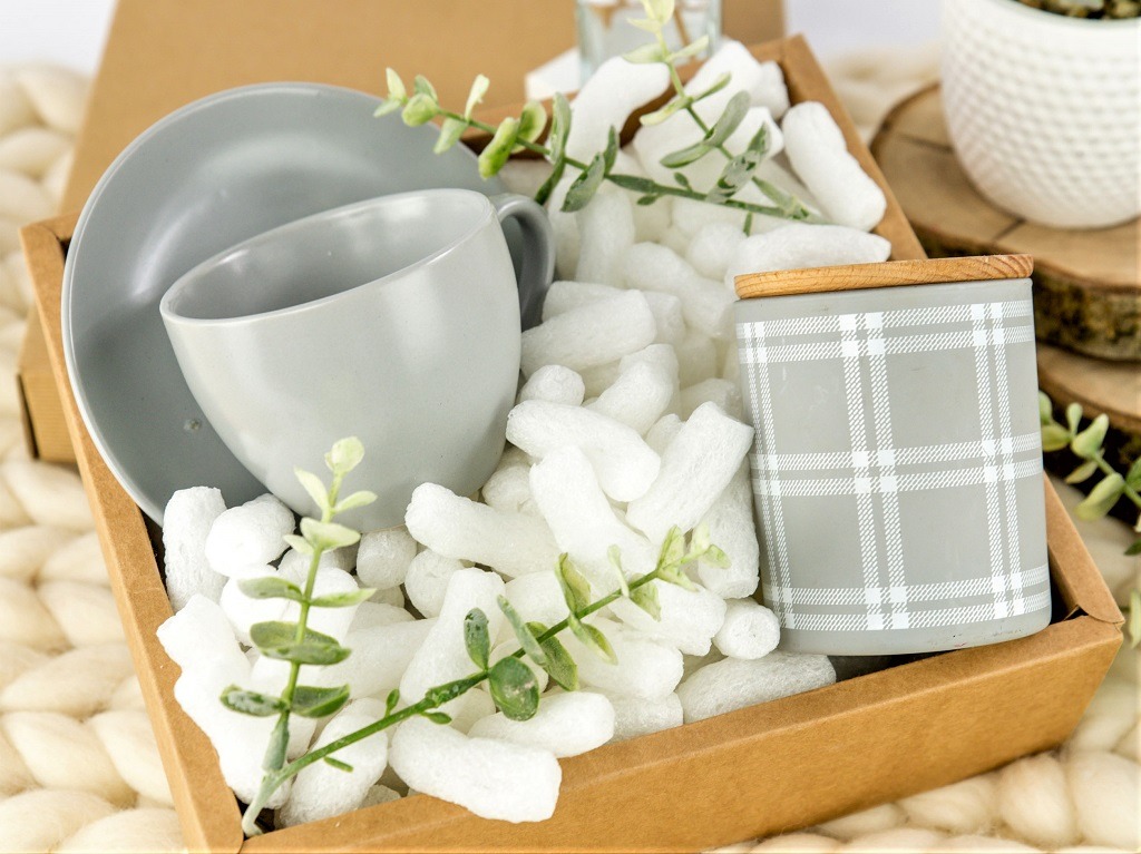 Szary zestaw filiżanki i talerzyka oraz świeca w pudełku wypełnionym białym wypełniaczem i zielonymi gałązkami.