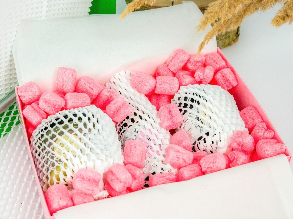 Różowe piankowe wypełniacze i białe siatkowe owijki zabezpieczają szklane butelki w różowym pudełku.