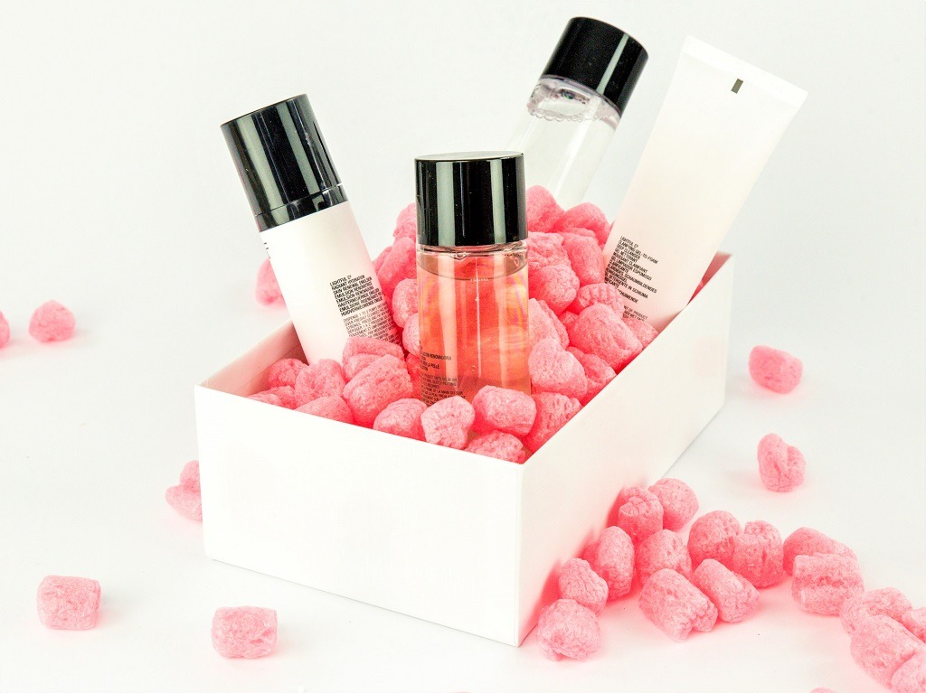 Białe pudełko wypełnione różowymi wypełniaczami, w którym umieszczono trzy kosmetyki w minimalistycznych opakowaniach: butelka z płynem, tuba i butelka z pompką.