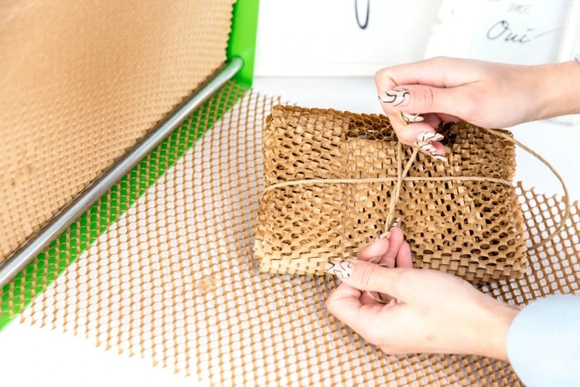 Owijanie prezentu brązowym papierem w kształcie plastra miodu i przewiązywanie sznurkiem, widoczne dłonie z wzorzystym manicure, fragment podajnika z zieloną obwódką.