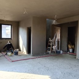 Mach BUD - Wnętrze w trakcie prac wykończeniowych: ściany pokryte świeżo nałożoną warstwą zaprawy, widoczny pracownik przy pracy, pętle grzewcze na podłodze, drabina, wiadra i narzędzia budowlane w tle.