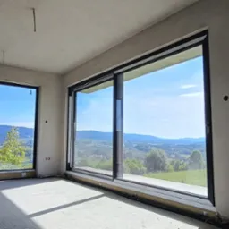 Surowe wnętrze z dwoma dużymi oknami przesuwnymi, przez które widać malowniczy krajobraz wzgórz i lasów. Ściany przygotowane do wykończenia, podłoga surowa.