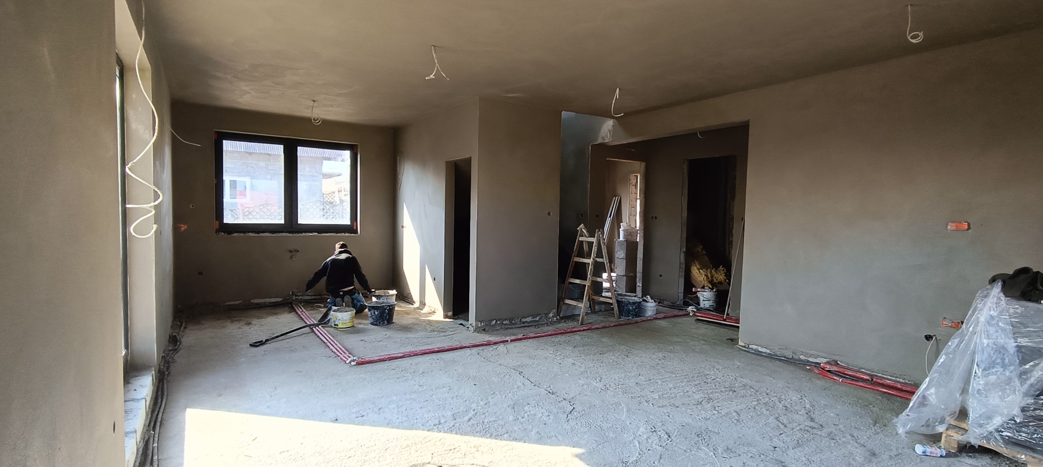 Wnętrze w trakcie prac wykończeniowych: ściany pokryte świeżo nałożoną warstwą zaprawy, widoczny pracownik przy pracy, pętle grzewcze na podłodze, drabina, wiadra i narzędzia budowlane w tle.